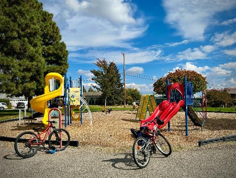 Photo of the play ground at Barranca Mesa Park. 2 bicycles parked in front of the entrance. Summer day  with green trees and fluffy clouds in the background.