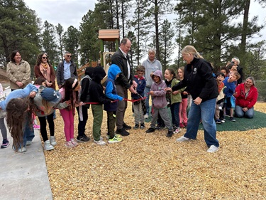 A man with a group of kids holds a large pair of scissors and cuts a red ribbon.