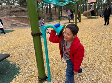 A kid in a red coat listens to another kid talking through a speaking and listening toy.
