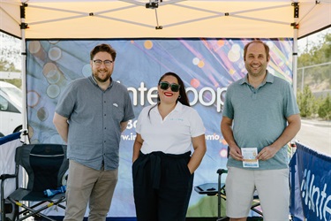 Three people stand under a shade tent.