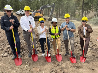 Six people stand with red shovels in a dirt field.