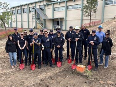 A large group of people stand with red shovels in a dirt field.