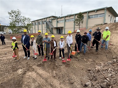A large group of people stand with red shovels in a dirt field.