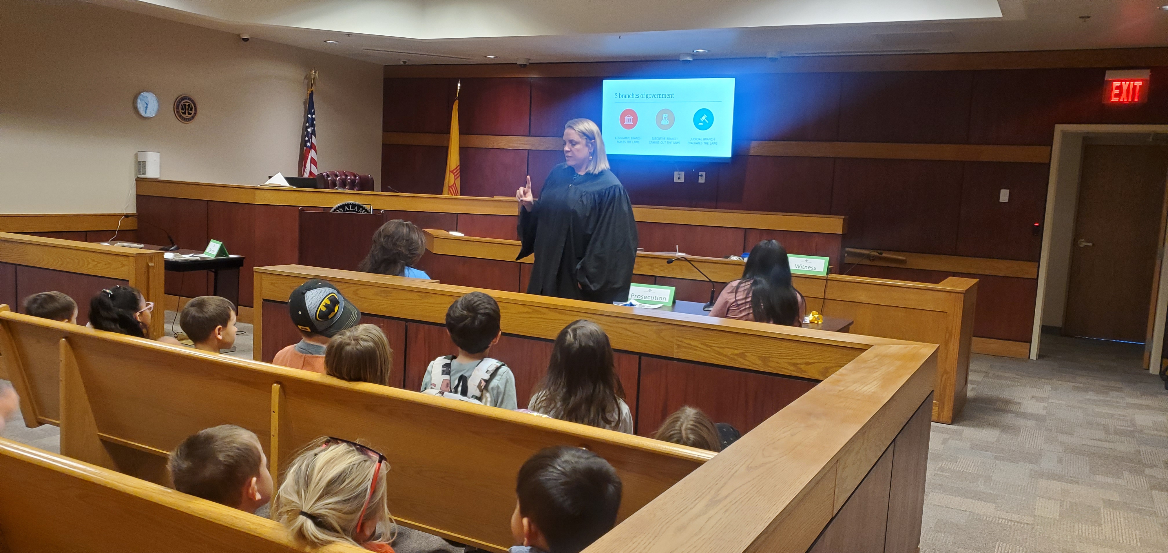 A group of children sit on benches while a judge in black robes talks to them