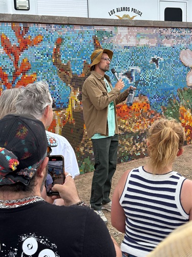 A man standing and talks into a microphone in front of a large mosaic wall.