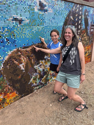 A woman and girl point at the top of a bear within a mosaic wall.