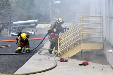 Two firefighters in bunker gear handle a hose preparing to enter a smoke filled building in a training exercise