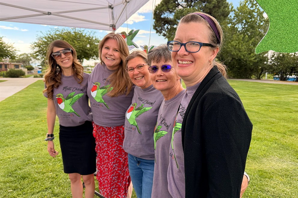 Photo of five social services staff members at Los Alamos County Farmers Market, standing together and smiling. All the staff are wearing matching shirts featuring their spokes-animal which is a hummingbird. 