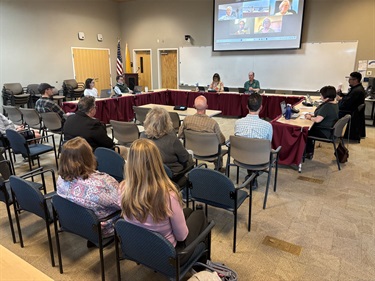 A group of people are seated at tables in a U shape in front of an audience.