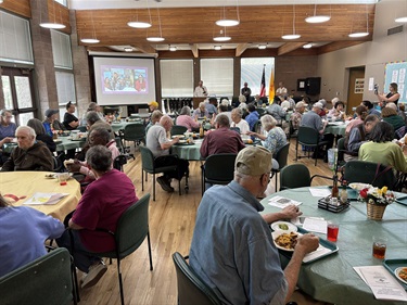 A large group of people eat lunch while a person speaks.
