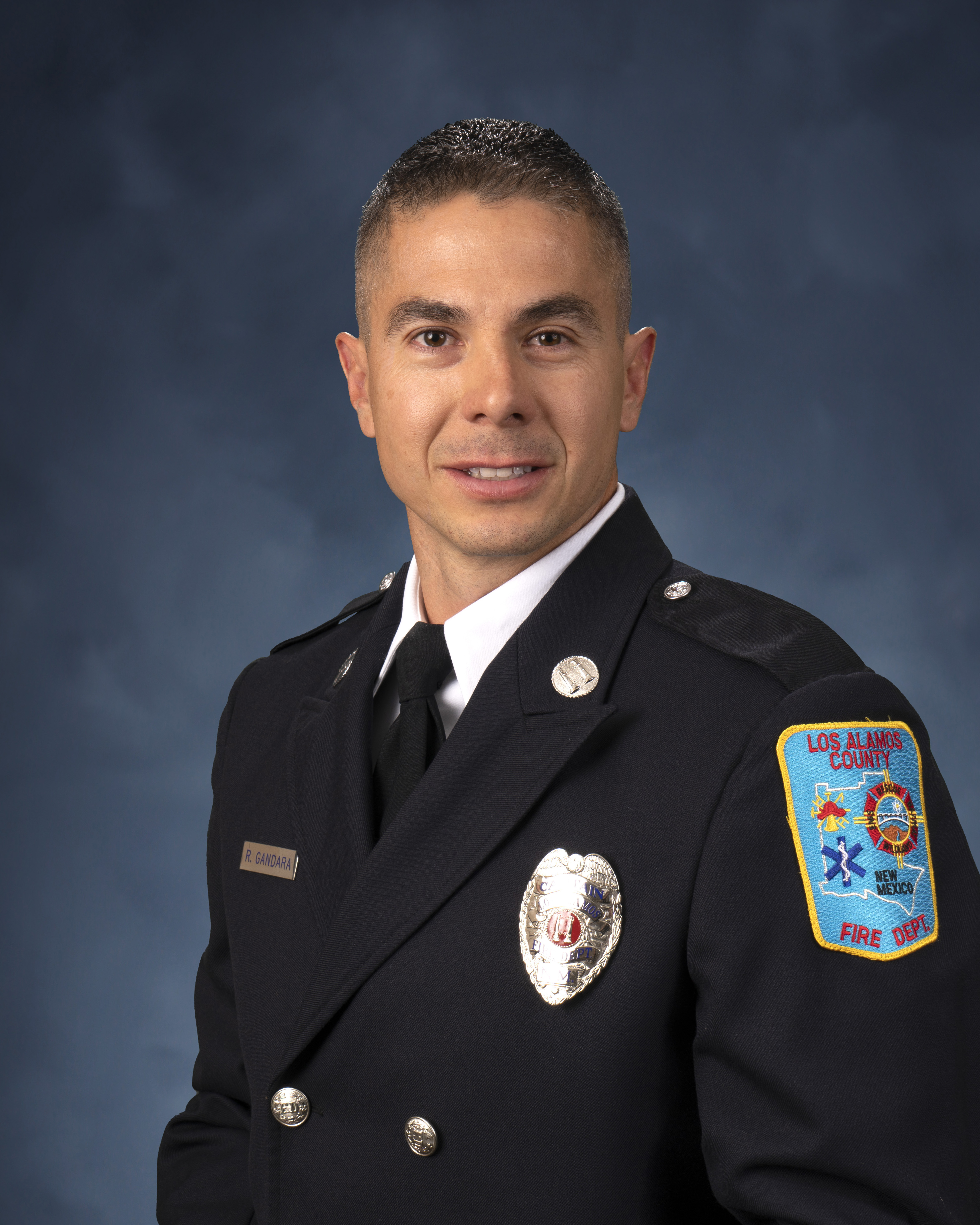 A portrait of a man in a formal suit with a metal badge on the front and a fire department patch on the shoulder.