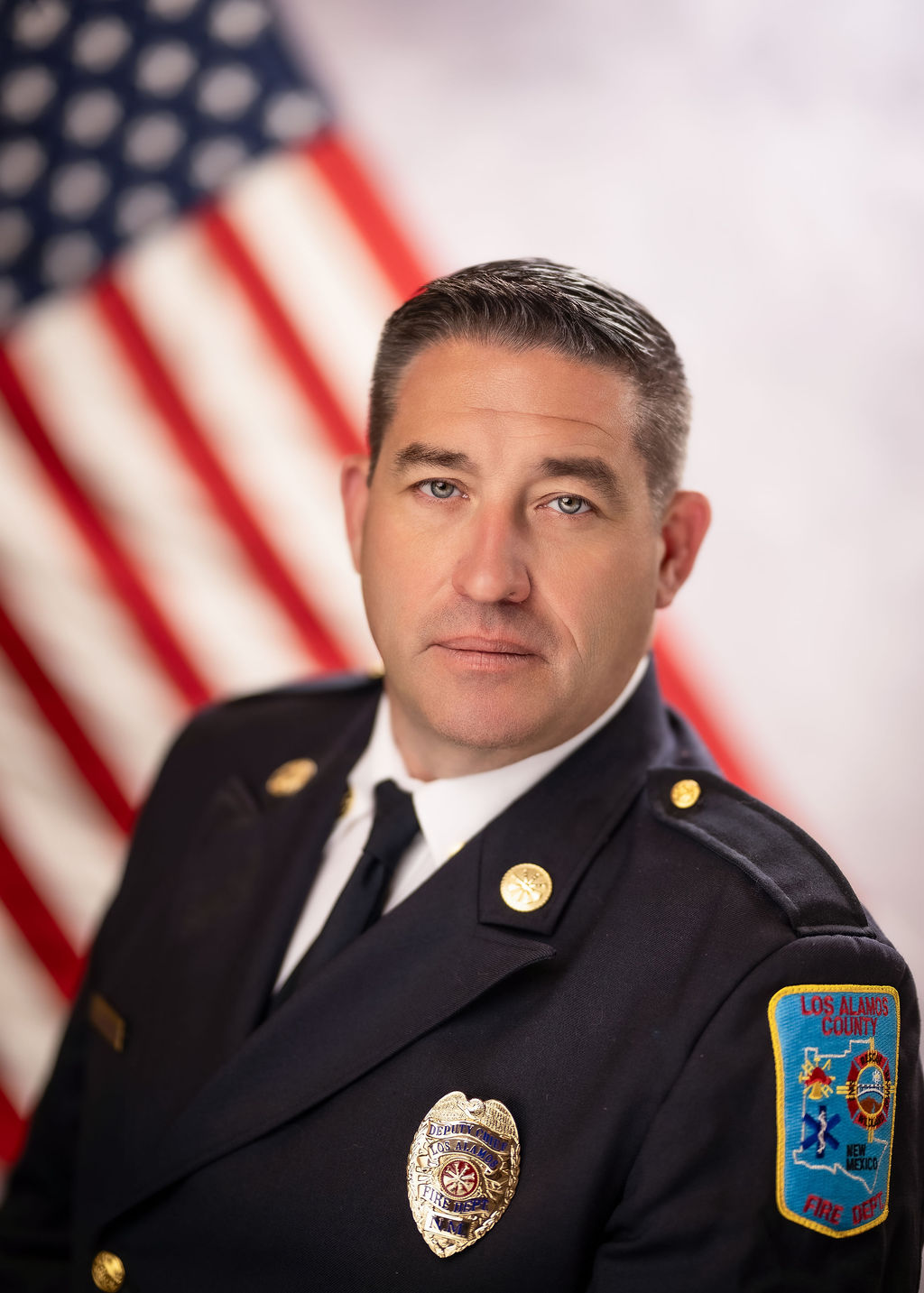 A portrait of a man in a formal suit with a metal badge on the front and a fire department patch on the shoulder.